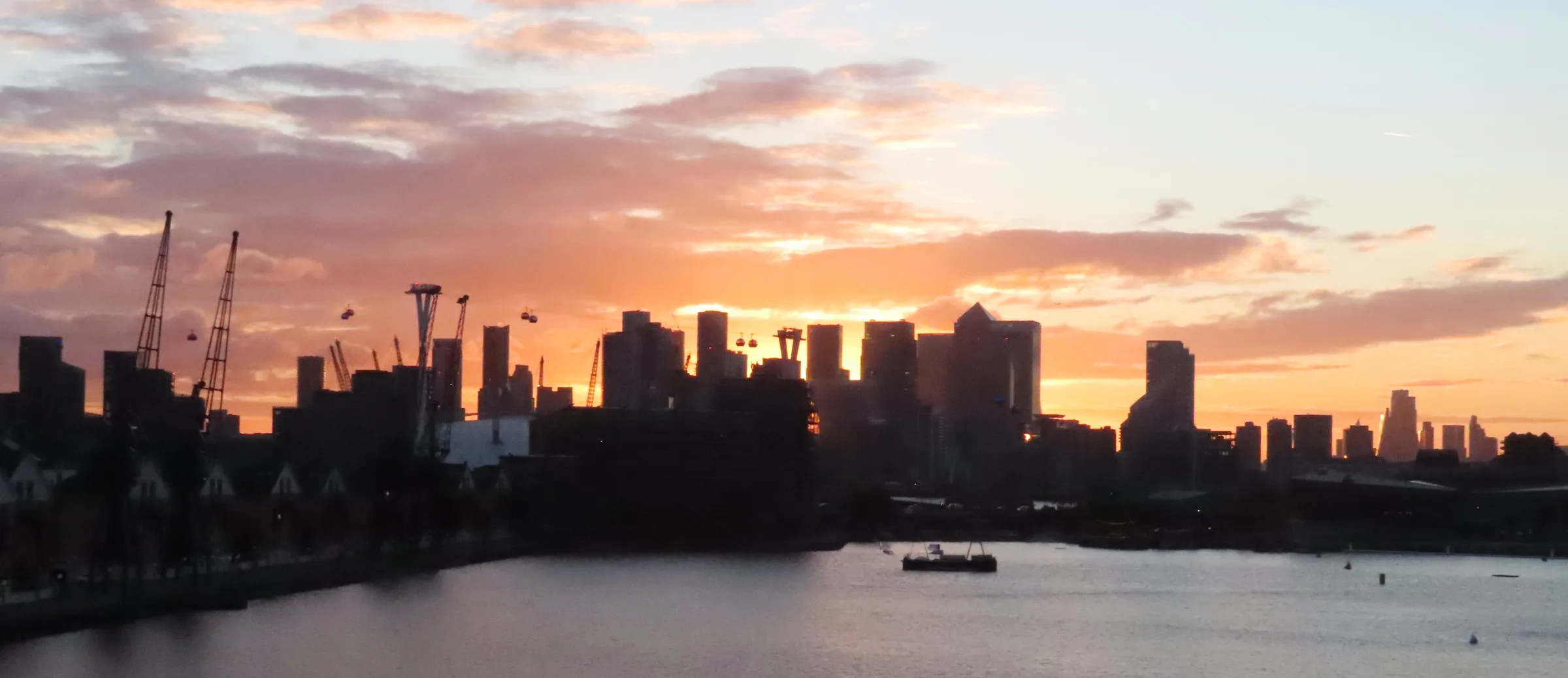 Open water at Royal Victoria Dock at sunset, with dockside cranes, the cable car and Canary Wharf beyond. Photograph taken in October 2024.